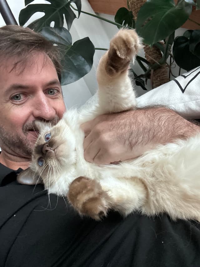 A man lying down with a fluffy cat resting on his chest, surrounded by green plants